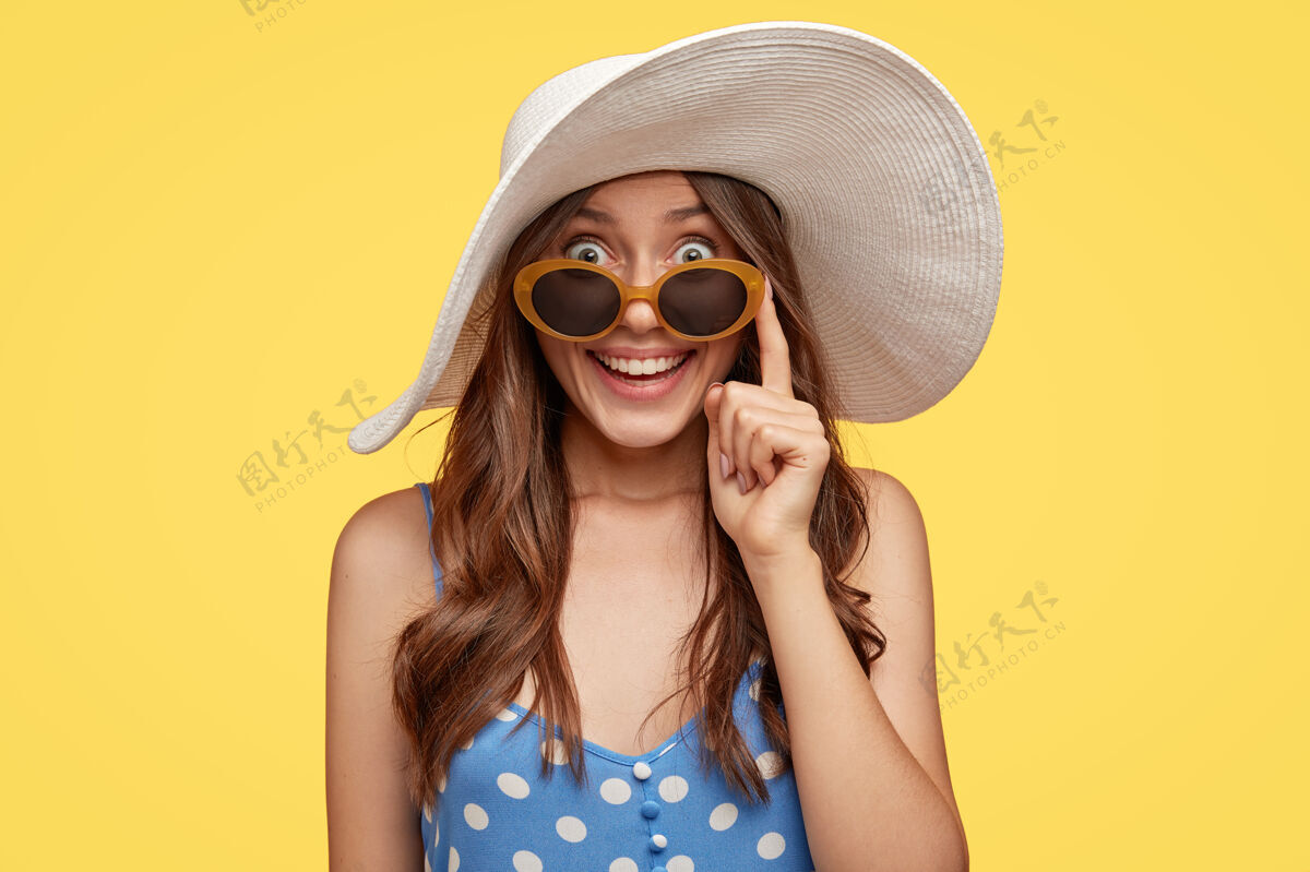 娱乐表情欢快的时尚女士 戴着白帽子和太阳镜 在度假期间找到酒店住宿 准备去海滩 隔着黄墙旅游和夏季概念女性化外表微笑