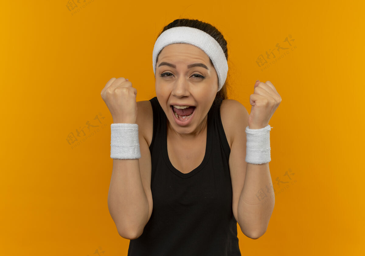 快乐身穿运动服 头箍紧握拳头的年轻健身女士站在橙色的墙上 既高兴又兴奋拳头运动员健康