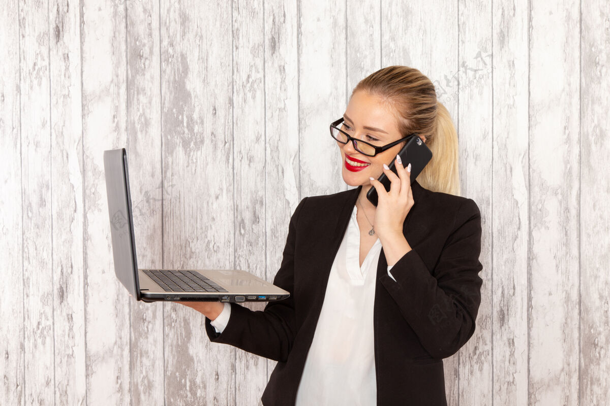 电话正面图：穿着黑色夹克的年轻女商人在白色办公桌上用笔记本电脑和电话交谈工作衣服严格