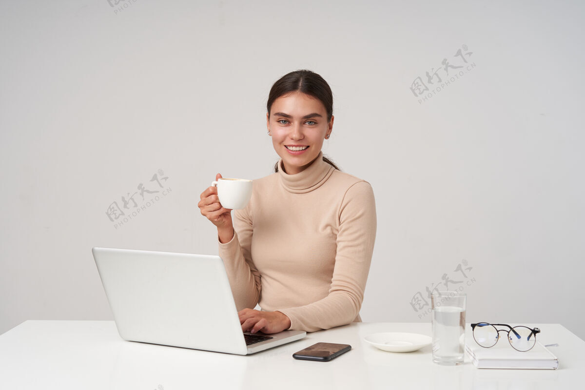 坐着年轻貌美的正派黑发女士 身着正装 坐在桌旁 拿着现代化的笔记本电脑 喝着咖啡 脸上洋溢着迷人的笑容Poloneck杯子商务女性