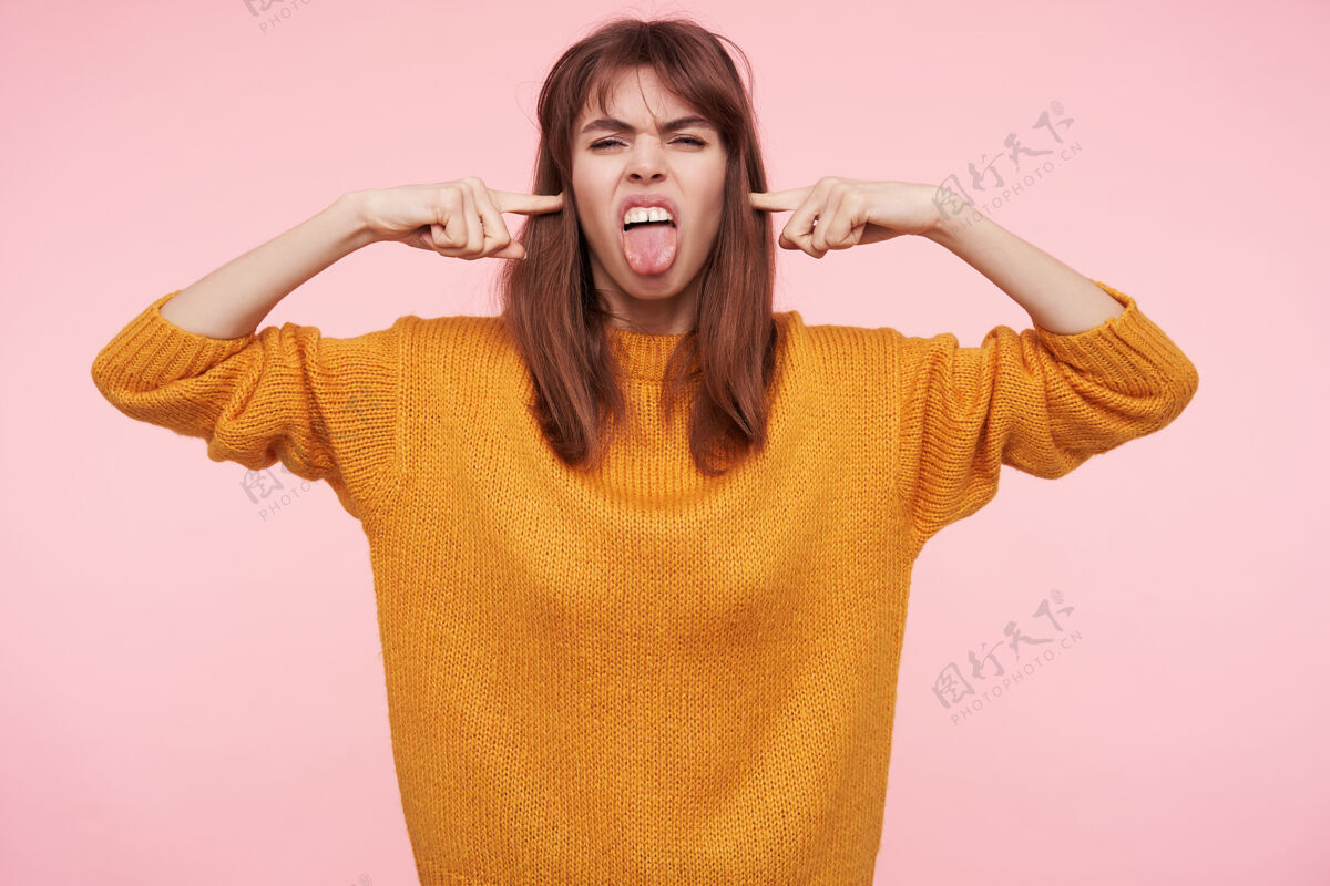 女士一张年轻的黑发女子的横幅照片 随意的发型在脸上做鬼脸 伸出舌头 把食指放在耳朵里 隔离在粉红色的墙上肖像头发衣服