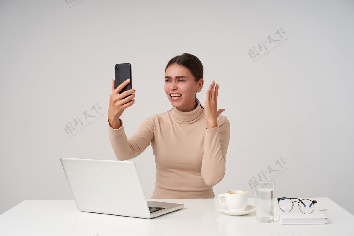 电话年轻迷人的黑发女士一边用智能手机举手 一边用现代笔记本电脑在办公室进行激动人心的电话交谈现代女士心情