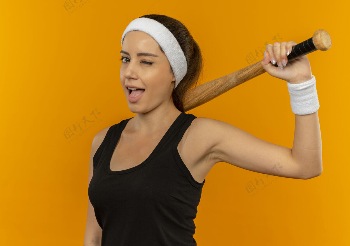 运动员身穿运动服 头箍 手持球棒 微笑着站在橙色墙壁上的年轻健身女士抱着运动装姿势