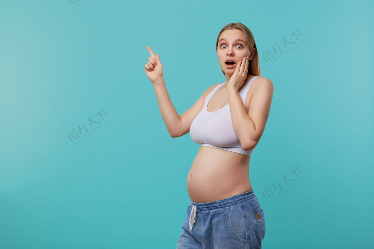 肖像惊呆了年轻的蓝眼睛金发长发女子举行了她的脸颊上抬起的手掌 同时看着相机 怀孕时 在蓝色的背景摆姿势自然女士未来