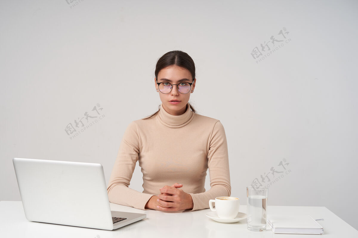 欧式严肃的年轻黑发女商人戴着眼镜 穿着正式的衣服 坐在桌旁拿着笔记本电脑 双手折叠在台面上 在白色的墙上摆姿势20多岁姿势发型