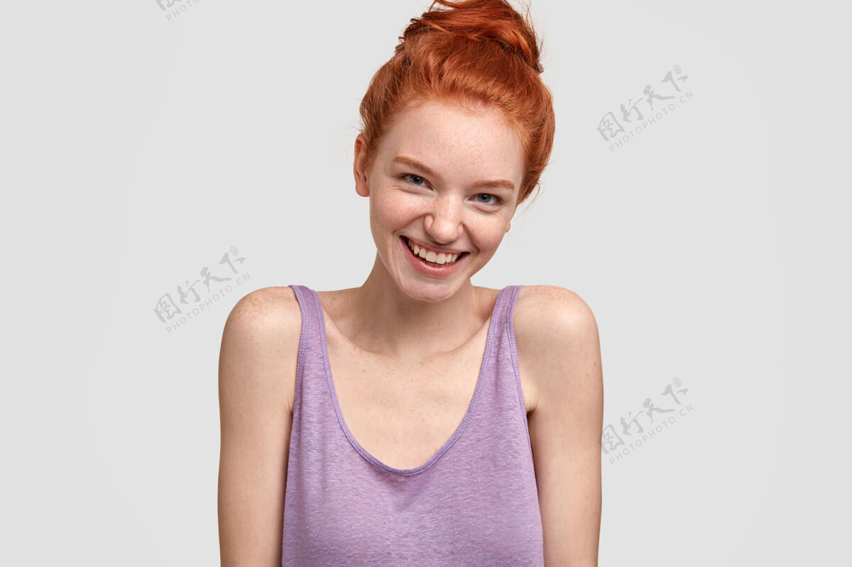 表情照片中的友好交际的女人有着红发和完美的雀斑皮肤 愉快地凝视着 有着温柔的微笑 穿着休闲的超大背心 露肩 模特靠着白墙满意女人站