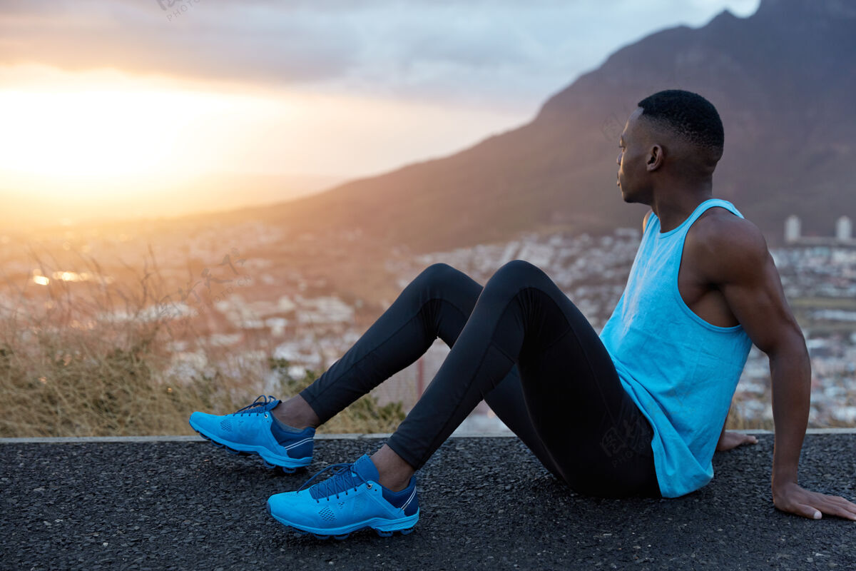 民族健美男坐在一边 皮肤黝黑 手肌肉发达 穿着运动服 注视着日出 在山上摆姿势 跑步后休息运动 自然运动员激烈精力充沛