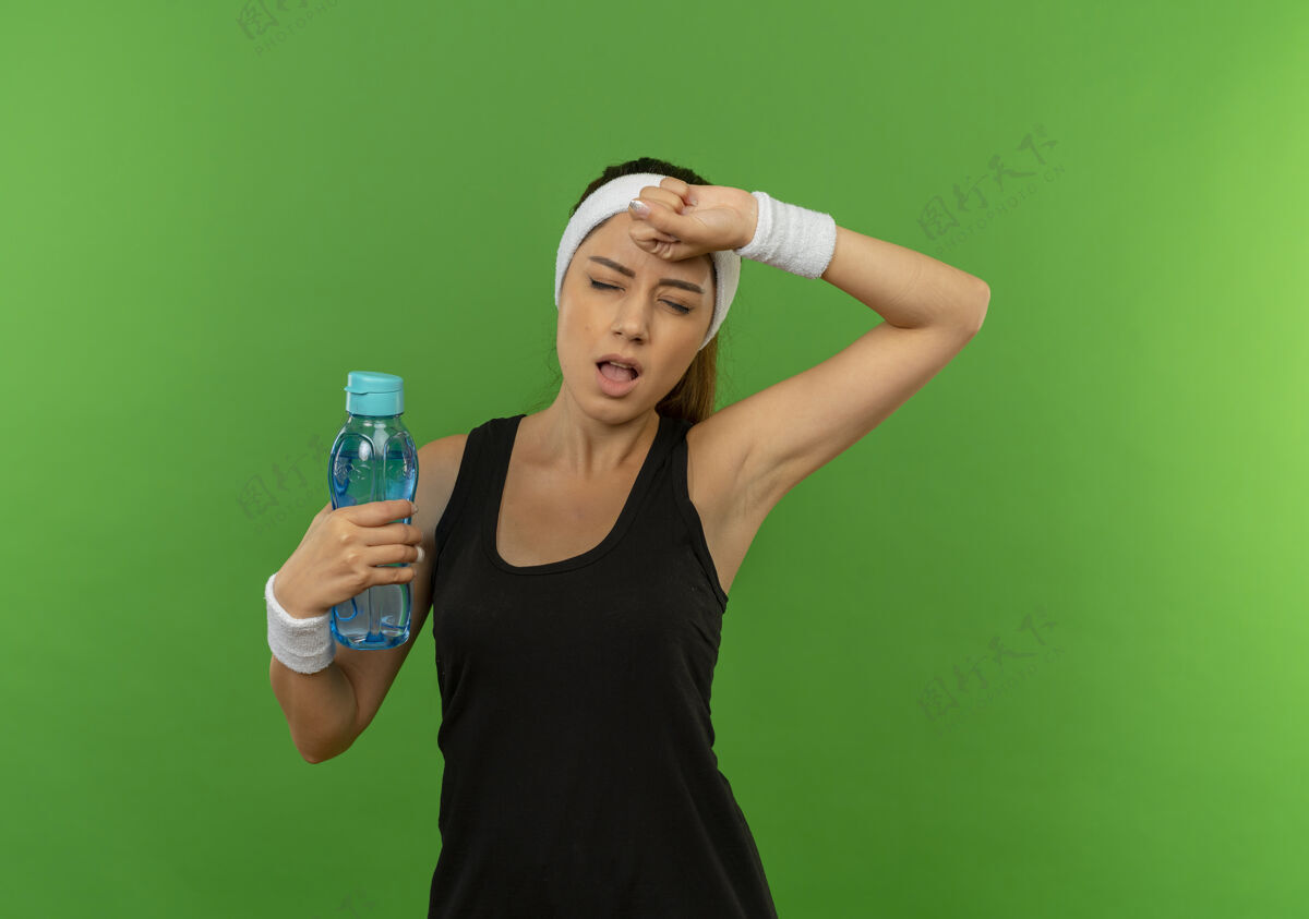运动穿着运动服的年轻健身女 头上戴着头巾 手里拿着一瓶水 站在绿色的墙上 看上去又累又累筋疲力尽看着人