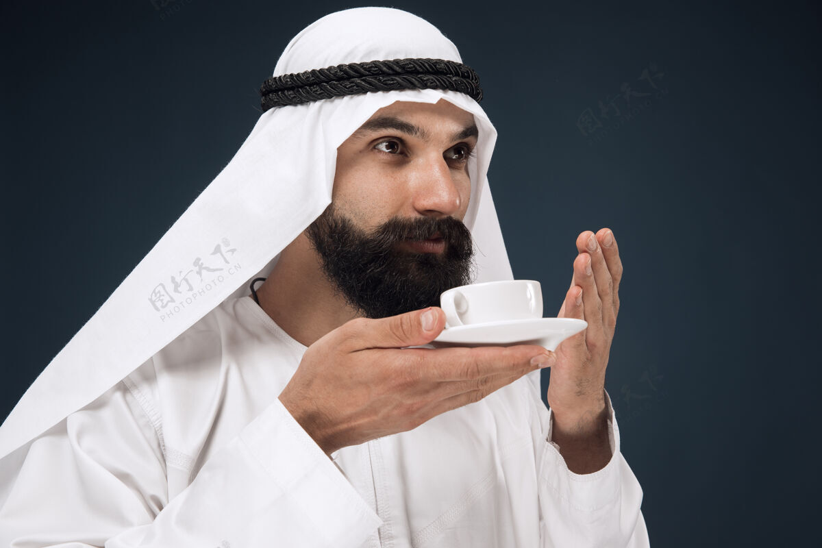 围巾深蓝色工作室背景上的半身阿拉伯沙特商人肖像年轻男模站着喝咖啡或茶商业 金融 面部表情 人类情感的概念家伙反应微笑