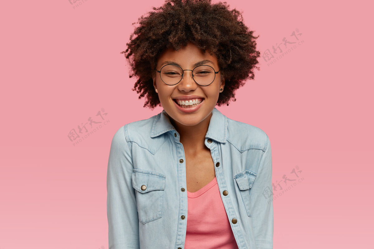 室内积极的女人 健康的皮肤 灿烂的笑容 享受生活 听到谈话者的好消息后心情愉快 戴着透明的眼镜 隔着粉红色的墙魅力牙齿眼镜