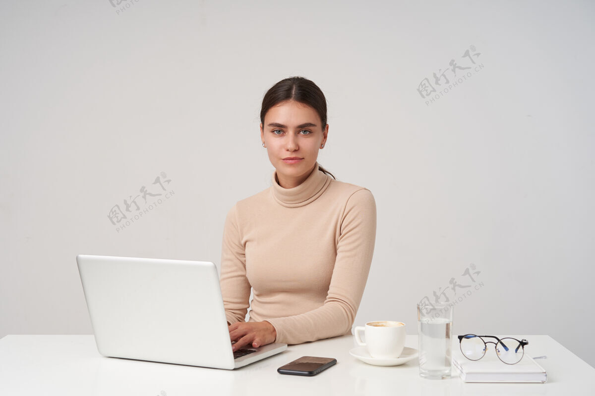 优雅迷人的年轻黑发女商人在现代办公室里工作 手里拿着笔记本电脑 双手放在键盘上 积极地看着摄像机 隔着白墙Poloneck正式玻璃