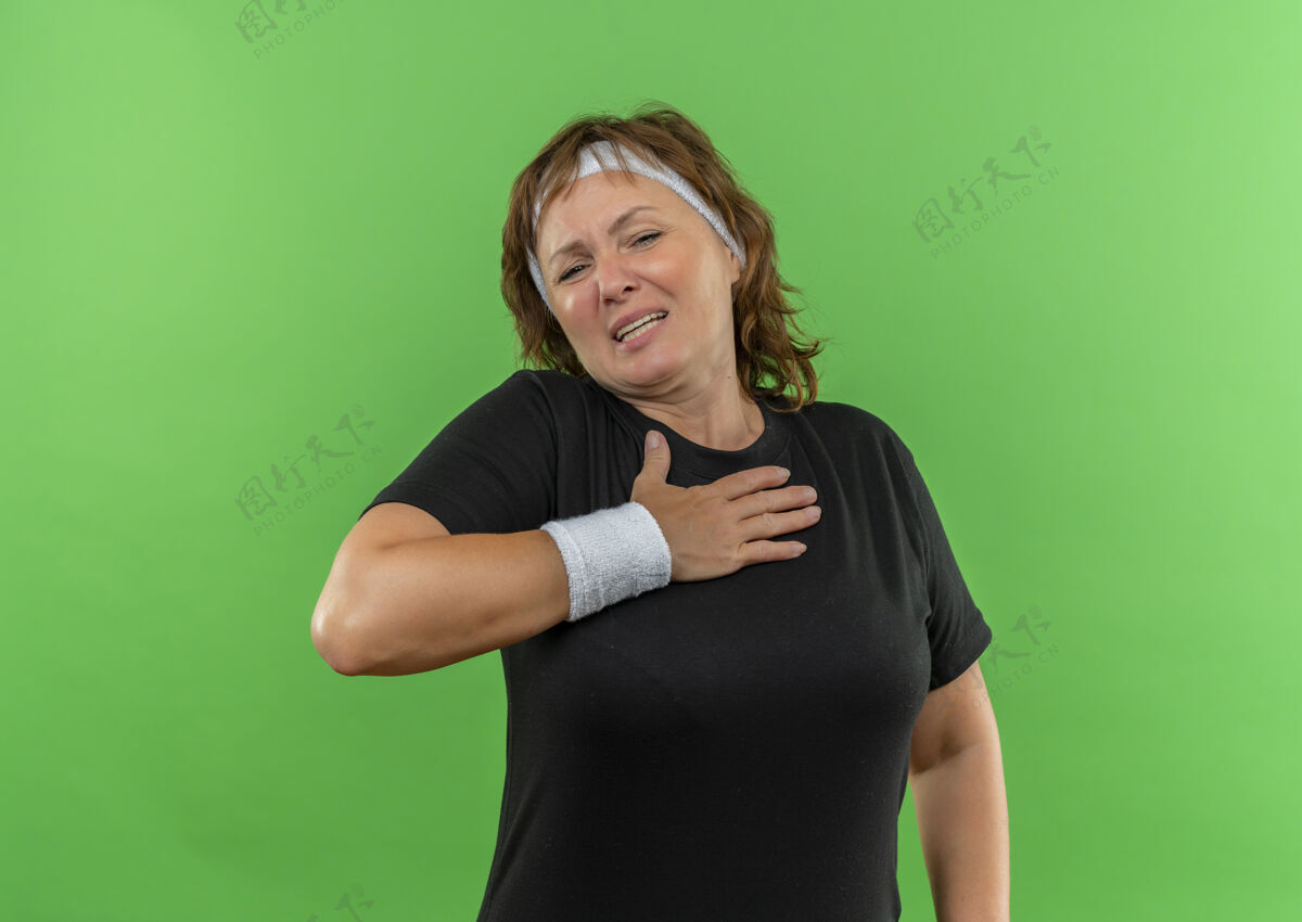 运动装身穿黑色t恤的中年运动女性 头箍手放在胸前 站在绿色的墙上 看上去很不舒服中间年龄人