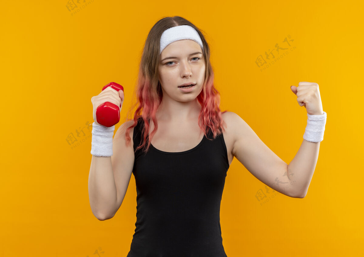 站穿着运动服的年轻健身女士举着哑铃举起拳头 像一个胜利者站在橙色的墙上健身年轻人