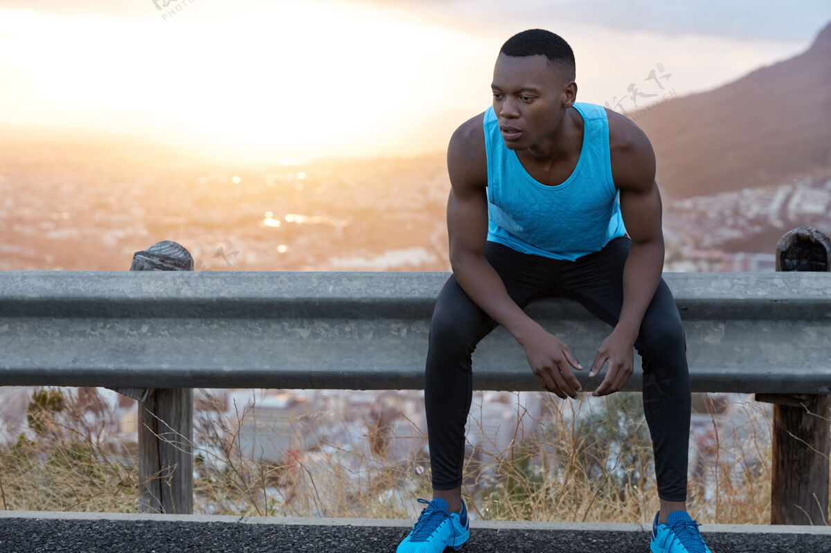 运动疲惫的非洲裔美籍男子形象 表情沉思 目光低垂 紧张训练后感到疲惫 坐在路标旁 美丽的日出与信息复制空间疲劳男人运动鞋
