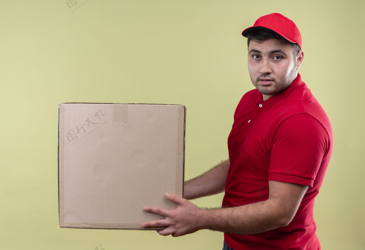 人年轻的送货员穿着红色制服 戴着帽子 手里拿着纸箱 自信地站在绿色的墙上年轻人纸板帽子
