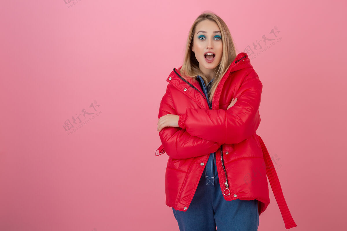 外观金发碧眼的活跃女性穿着色彩鲜艳的红色冬季羽绒服在粉色墙面上摆造型 尽情玩乐 温暖的外套时尚潮流 惊喜震惊的表情天气服装女士