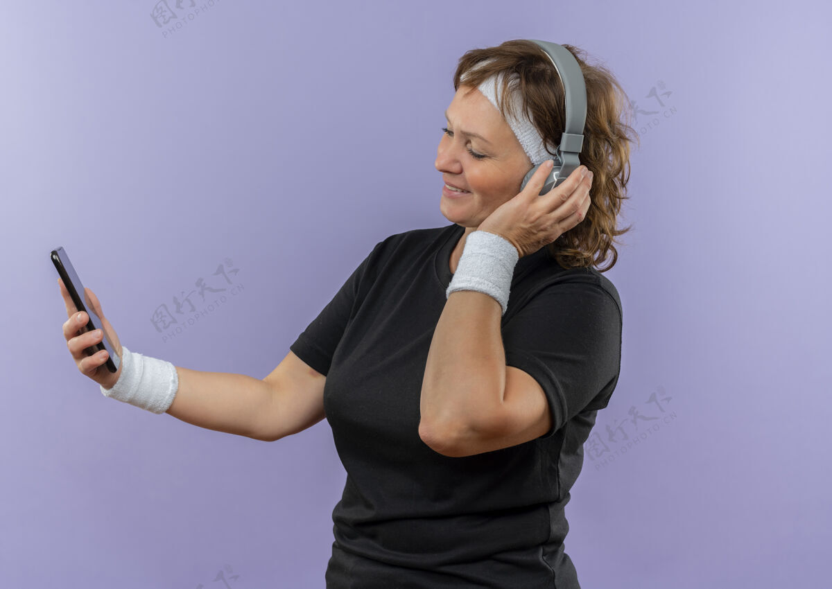 站立一位中年运动女性 身穿黑色t恤 戴着头带和耳机 站在蓝色的墙上 看着手机屏幕上搜索音乐音乐搜索耳机