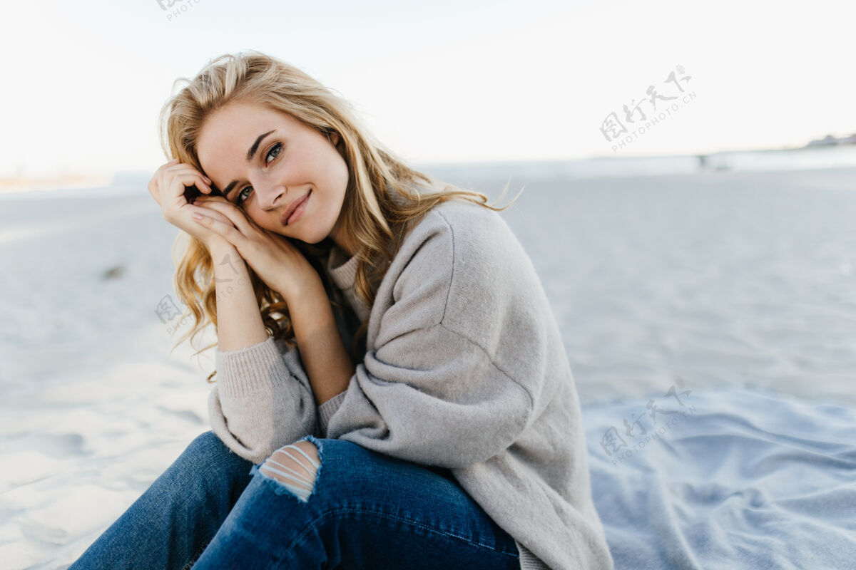 肖像穿着牛仔裤和毛衣的绿眼睛女人坐在沙滩上 跪在海里周末水放松