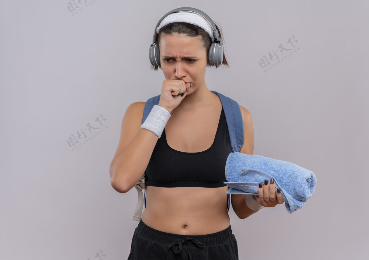 背包穿着运动服的年轻健身女士 背着背包 头上戴着耳机 拿着毛巾 用拳头靠近下巴 站在白墙上思考姿势靠边公民