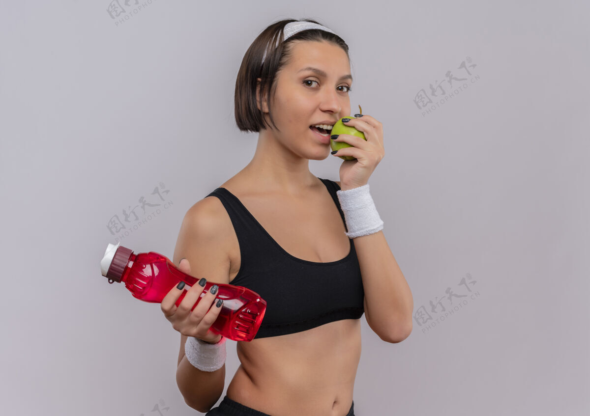 女性穿着运动服的年轻健身女士 手里拿着一瓶水和绿苹果 微笑着咬着苹果站在白色的墙上运动装站着健康