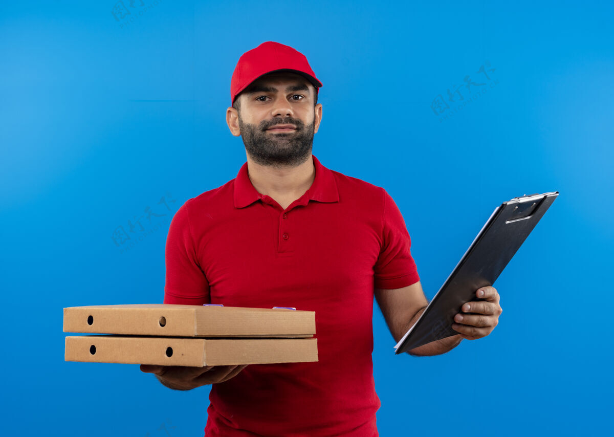 剪贴板满脸胡须 身穿红色制服 头戴鸭舌帽的送货员站在蓝色的墙上 手里拿着一叠披萨盒和剪贴板 表情自信胡子拿着送货