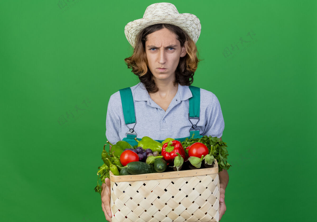 男人年轻的园丁穿着连体衣戴着帽子拿着装满新鲜蔬菜的箱子板条箱充分蔬菜