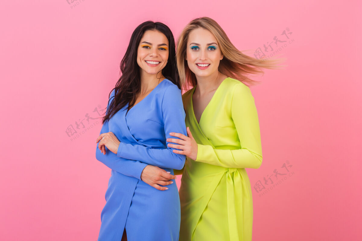 穿着两个时尚性感的微笑迷人的金发和黑发女子在粉红色的墙上摆姿势在蓝色和黄色的时尚多彩的礼服 夏季时尚潮流人奢侈季节