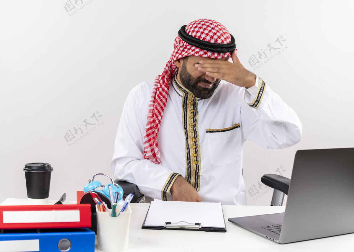 桌子身着传统服装的阿拉伯商人坐在桌旁 手提电脑看起来很疲惫 无聊地捂着眼睛在办公室工作笔记本电脑累穿