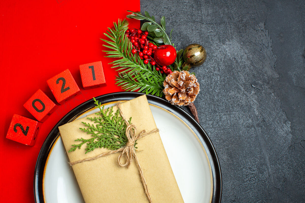 树枝新年背景的半张照片 餐盘上有礼物装饰配件 黑色桌子上的红餐巾上有杉木树枝和数字餐巾一半冷杉
