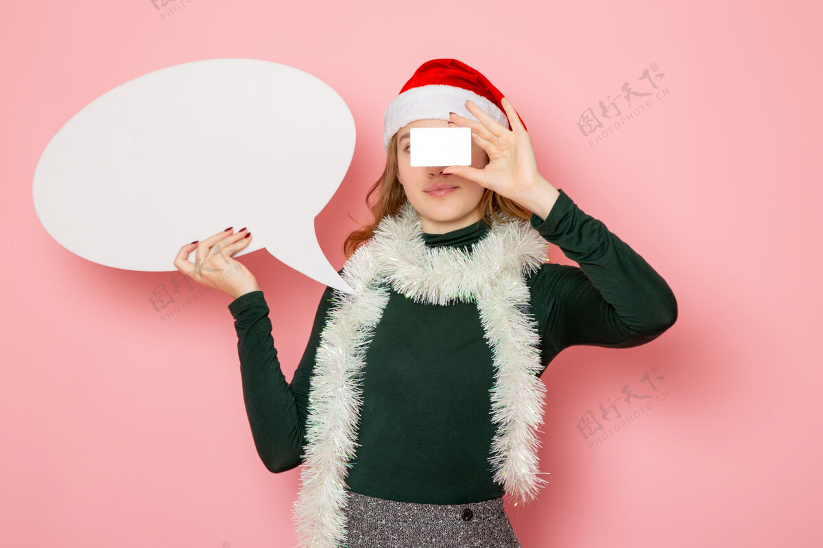 看法正面图年轻女性手持白色大牌子和银行卡在粉色墙上模型圣诞节服装肖像大