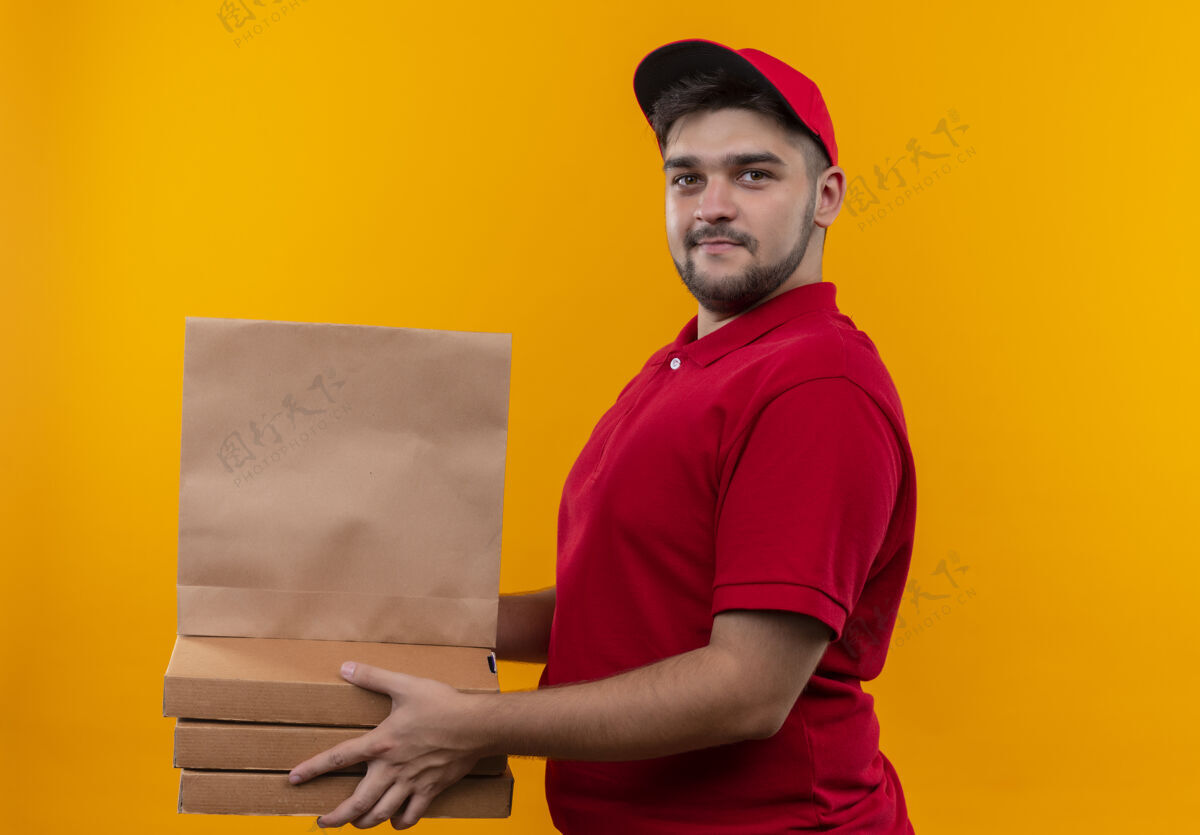 制服年轻的送货员穿着红色制服 戴着帽子 手里拿着纸包和一叠比萨饼盒 看上去很自信帽子叠着拿着