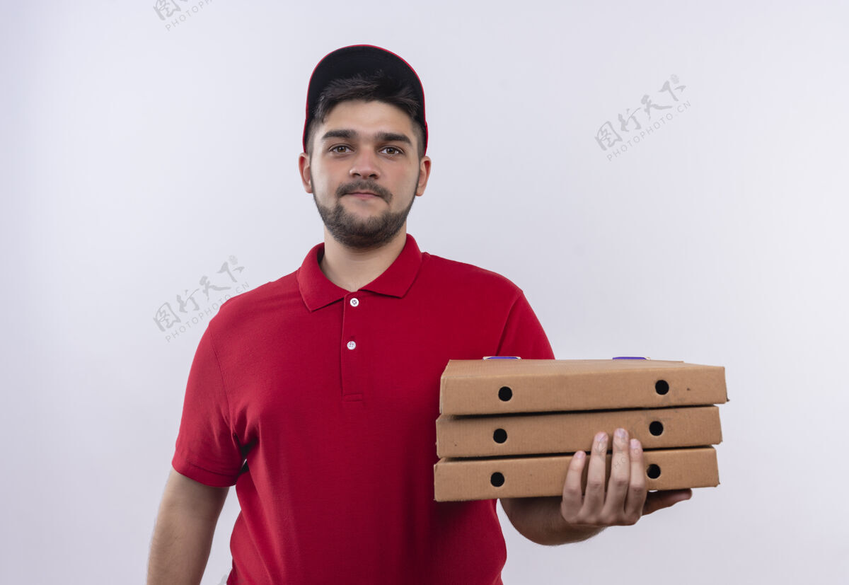 拿着年轻的送货员身穿红色制服 头戴鸭舌帽 手里拿着一叠披萨盒 看上去自信满面笑容看着红色帽子