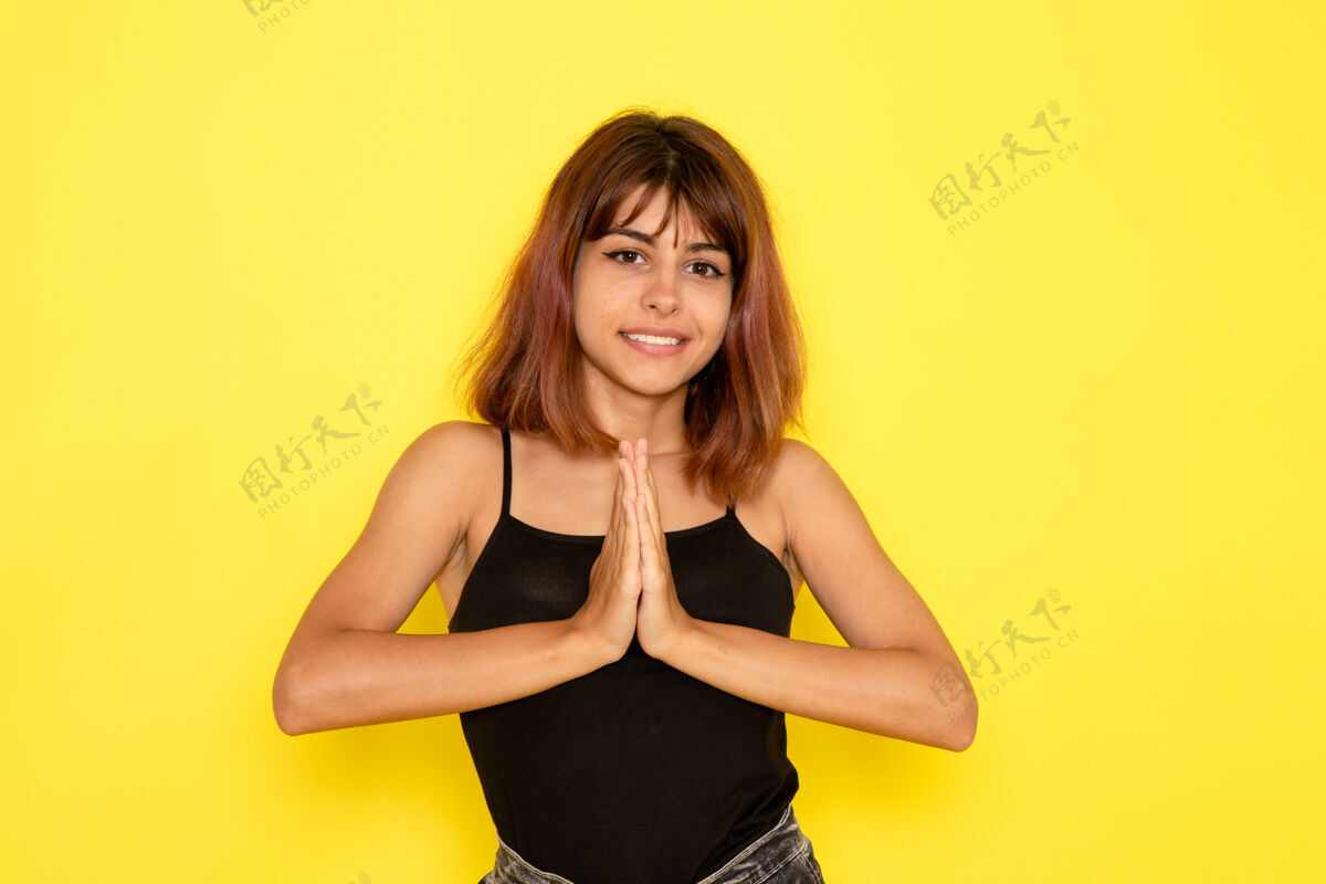 轻身穿黑色衬衫和灰色牛仔裤的年轻女性在浅黄色的墙上微笑着摆姿势姿势性感牛仔裤
