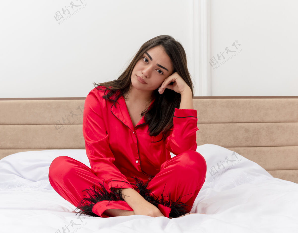 卧室穿着红色睡衣的年轻漂亮女人坐在床上看着摄像机 脸上带着悲伤的表情 在卧室内部的灯光背景下坐着床悲伤