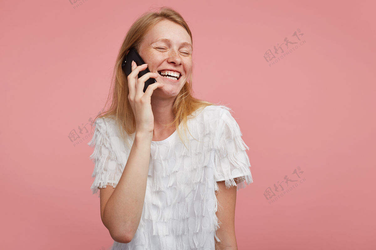 女性可爱的年轻快乐女人的肖像 浓密的头发 高举着智能手机 愉快地微笑 同时进行愉快的交谈 隔离在粉红色的背景下T恤手机年轻