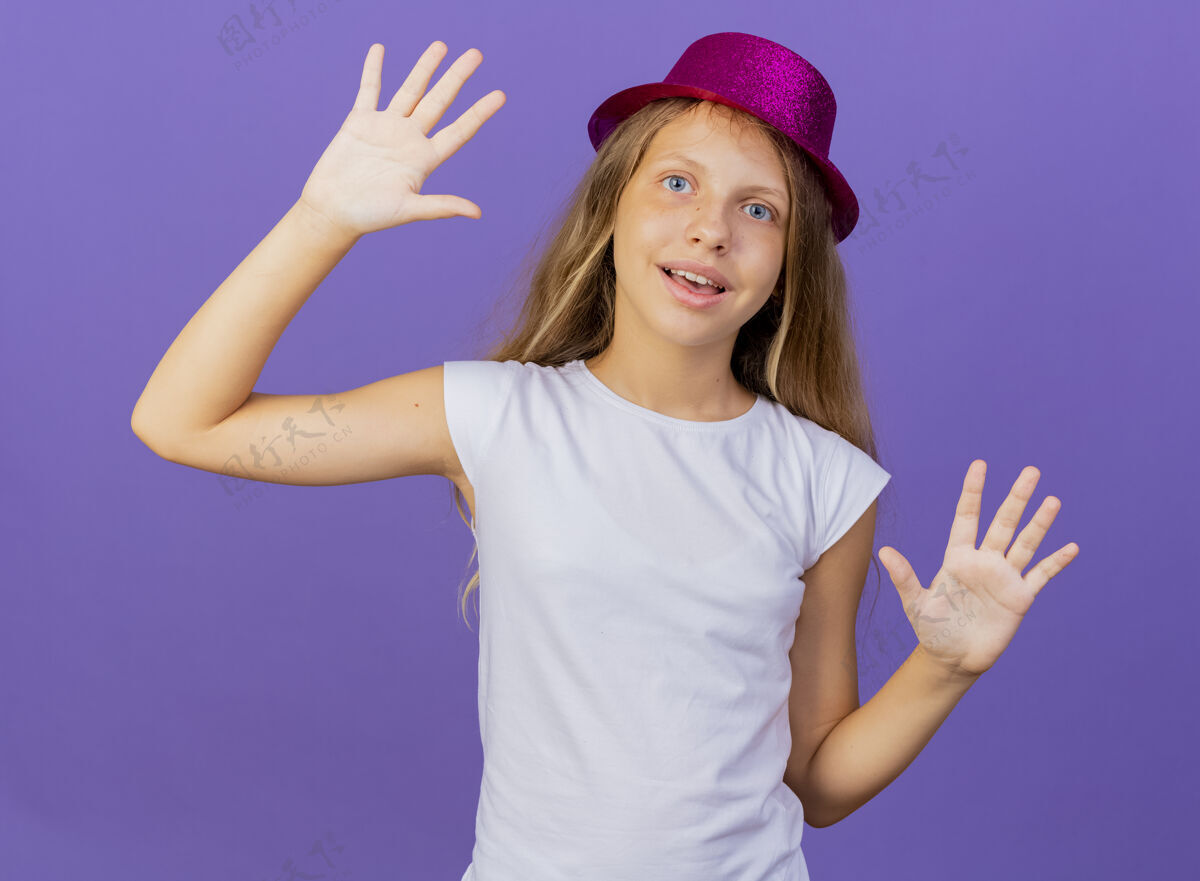 站着美丽的小女孩戴着节日帽子扬起手掌 开心而积极的微笑 生日派对的概念站在紫色的背景上快乐手掌节日