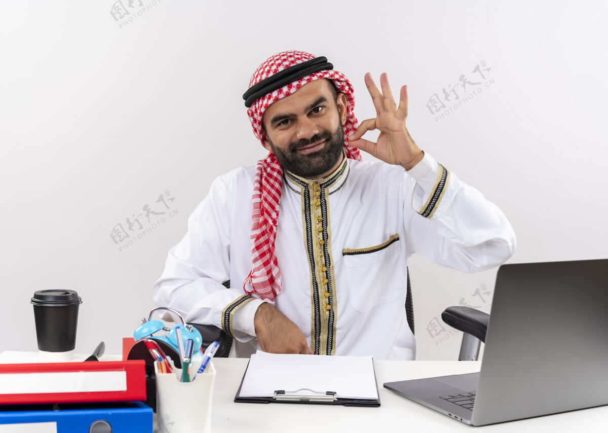桌子身着传统服饰的阿拉伯商人坐在桌旁 拿着笔记本电脑兴高采烈地做着ok标志在办公室工作穿商务人士电脑