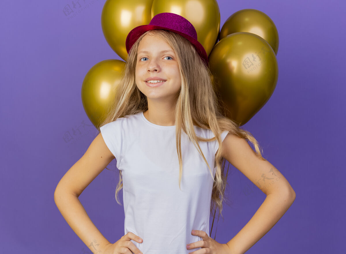 帽子戴着节日礼帽的漂亮小女孩和一群巴隆人看着相机开心地笑着 紫色背景下站着生日派对的概念女孩漂亮紫色