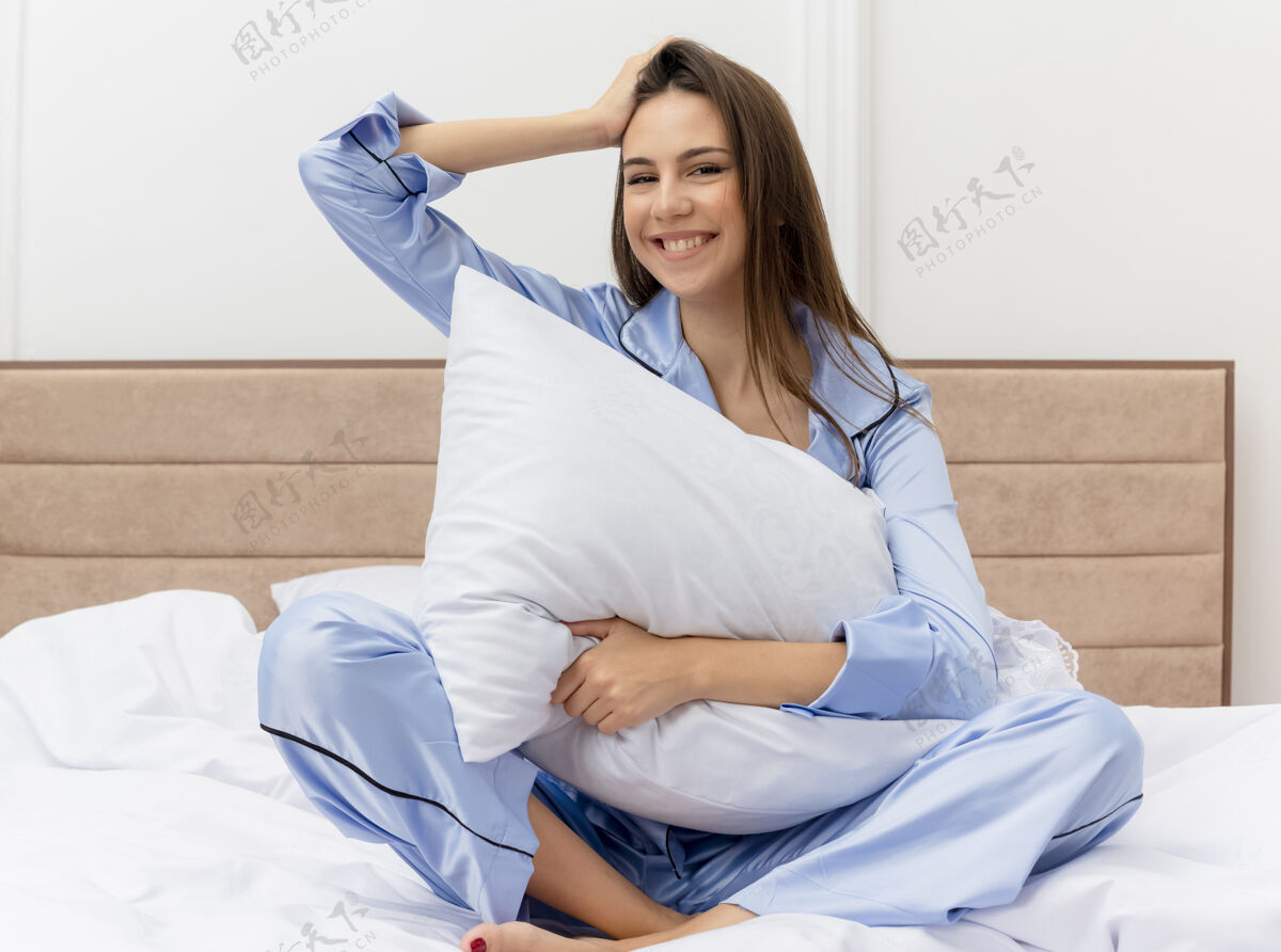 坐着穿着蓝色睡衣的年轻漂亮女人坐在床上 枕着枕头 看着相机 在灯光的背景下 她在卧室的内部笑容可掬年轻脸相机