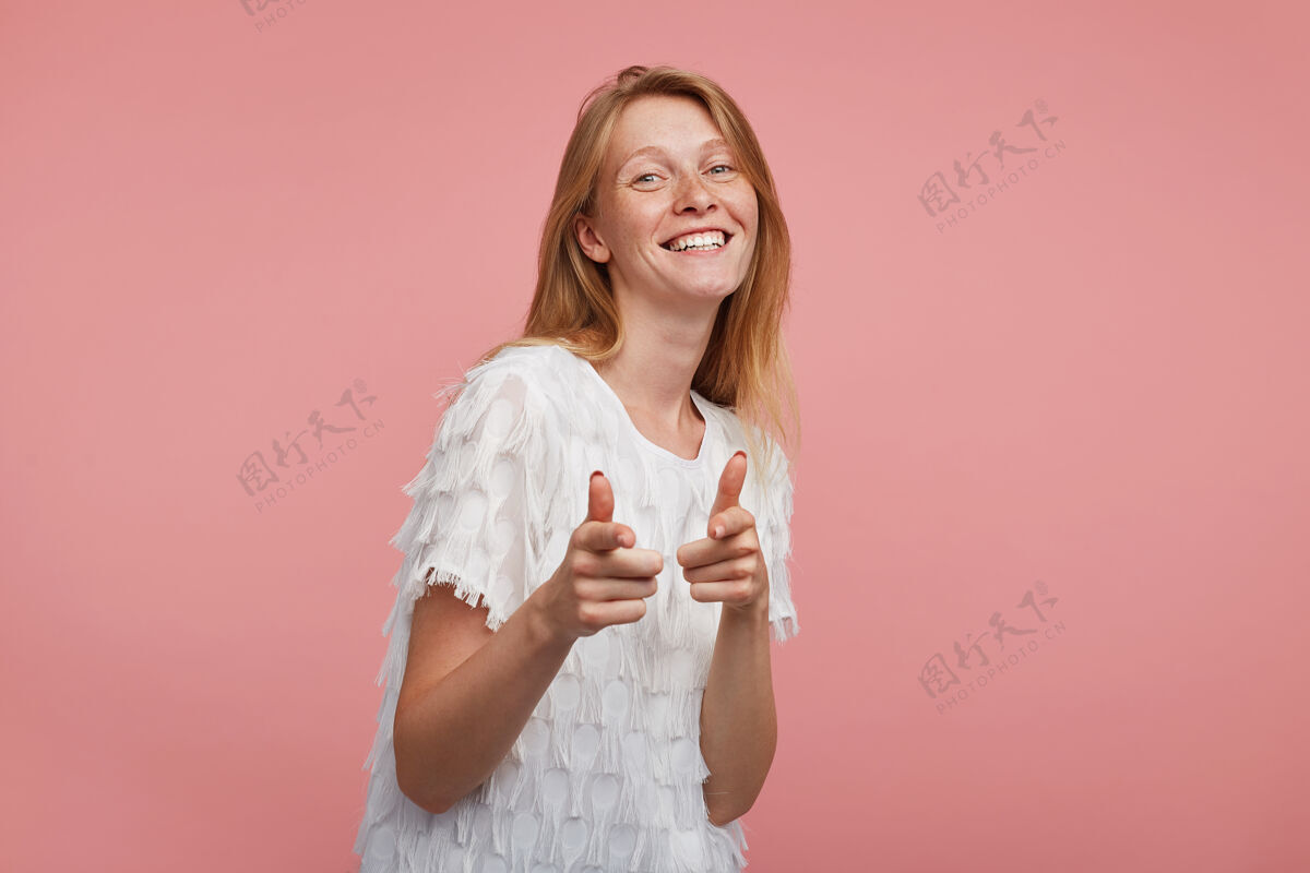 年轻室内拍摄的年轻快乐的红头发女人自然化妆指向相机与提高食指和微笑愉快 而在粉红色的背景摆姿势抬高微笑姿势