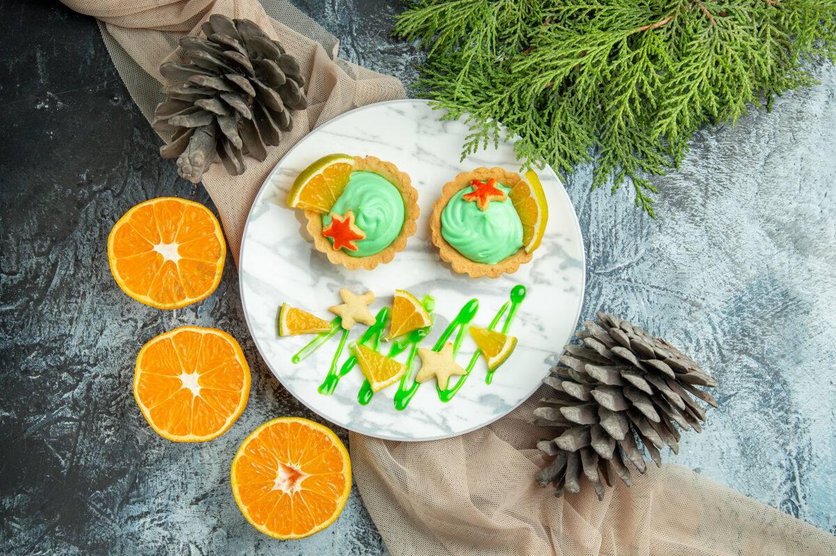 食物顶视图小馅饼与绿色糕点奶油和柠檬片在盘子上米色披肩切橘子松果在黑暗的桌子上馅饼盘子橙色