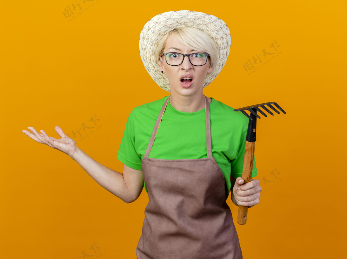 女人围裙上留着短发 戴着帽子 拿着小耙子的年轻园丁站在橙色的背景下 困惑而不高兴地看着摄像机看头发相机