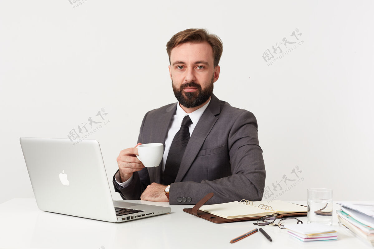 胡须照片中 年轻的留着胡须的黑发男性留着短发 面色平静地看着前方 一边用笔记本电脑和笔记本在白墙上工作 一边喝茶短嘴唇肖像