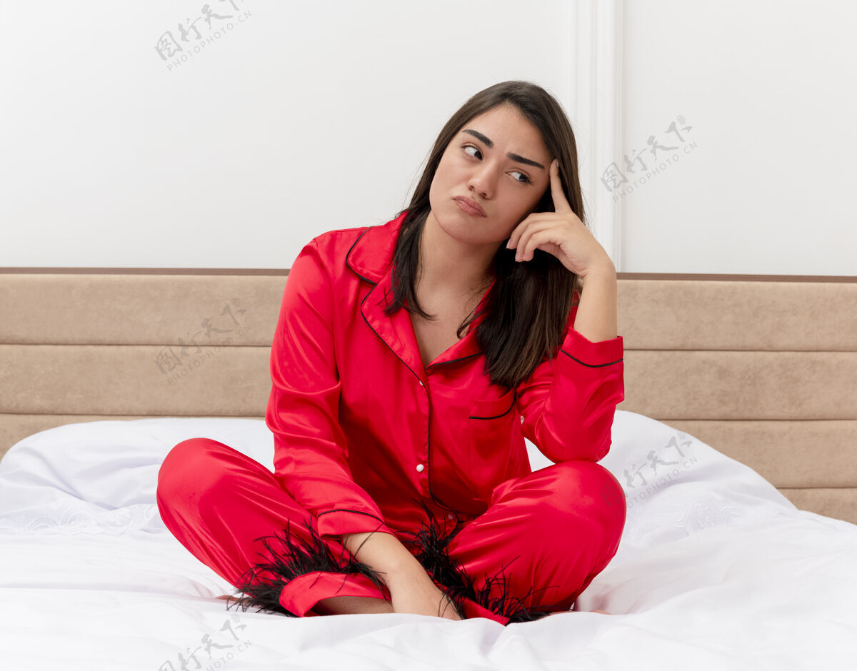 床穿着红色睡衣的年轻美女坐在床上 带着沉思的表情 在灯光背景下的卧室里看着不高兴红色漂亮卧室