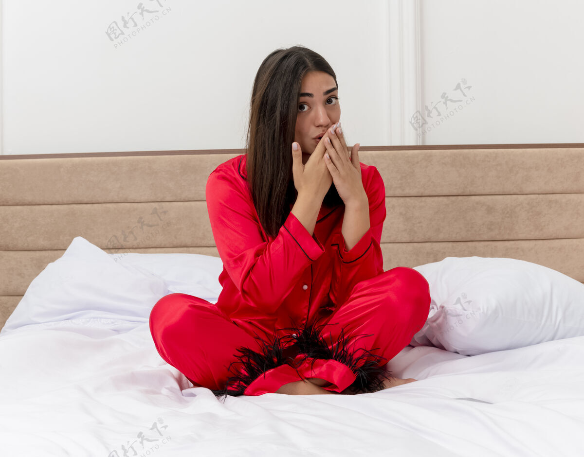 室内穿着红色睡衣的年轻美女坐在床上看着惊讶的凹嘴 双手放在卧室内部的灯光背景下床看着坐着