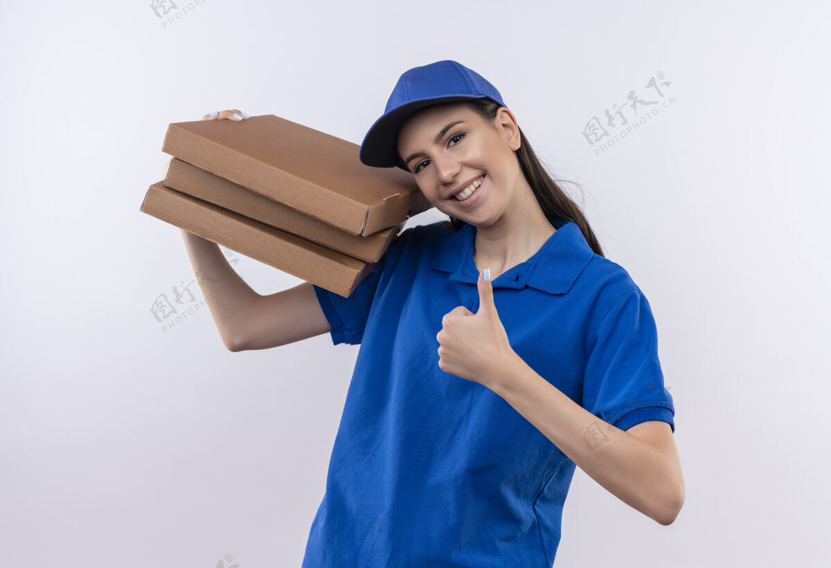 年轻身穿蓝色制服 头戴鸭舌帽的年轻送货女孩拿着一叠比萨饼盒 兴高采烈地微笑着看着镜头 竖起大拇指蓝色看微笑