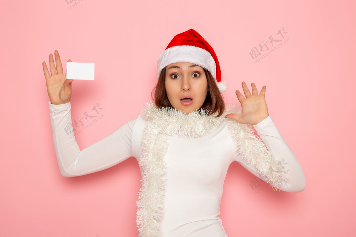肖像正面图年轻女性手持白色银行卡在粉色墙壁上圣诞节彩色节日新年时尚帽子银行色彩