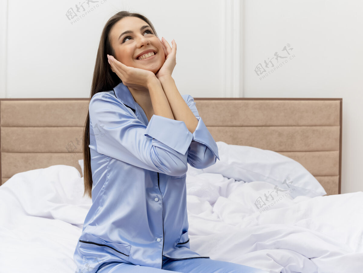 坐着穿着蓝色睡衣的年轻漂亮女人坐在床上仰视着 双臂放在脸上 在卧室的室内灯光背景下欢快地微笑着手臂睡衣室内