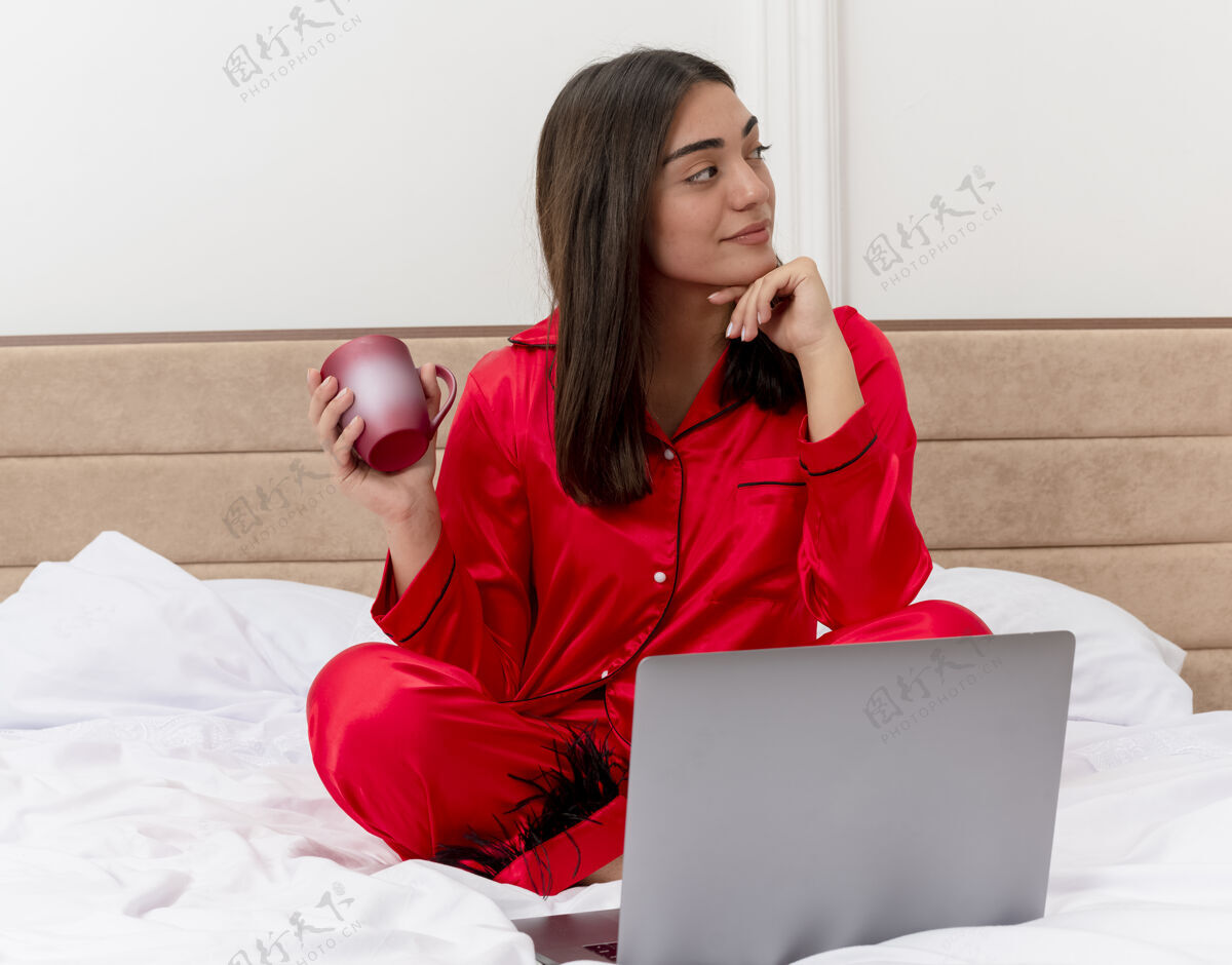 坐着穿着红色睡衣的年轻美女坐在床上 拿着笔记本电脑和一杯咖啡 一边看一边 脸上带着彭索夫的表情 在卧室内部的灯光背景下积极思考女人咖啡积极