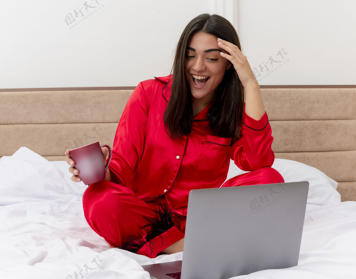 坐着穿着红色睡衣的年轻美女坐在床上 手里拿着笔记本电脑和一杯咖啡 在卧室的室内灯光背景下愉快而兴奋地微笑着睡衣杯子室内